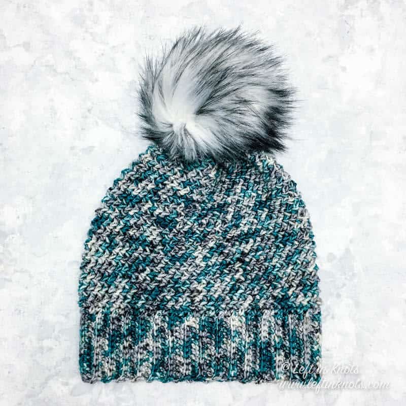 A blue and gray crochet beanie made with hand dyed yarn and a faux fur pom pom