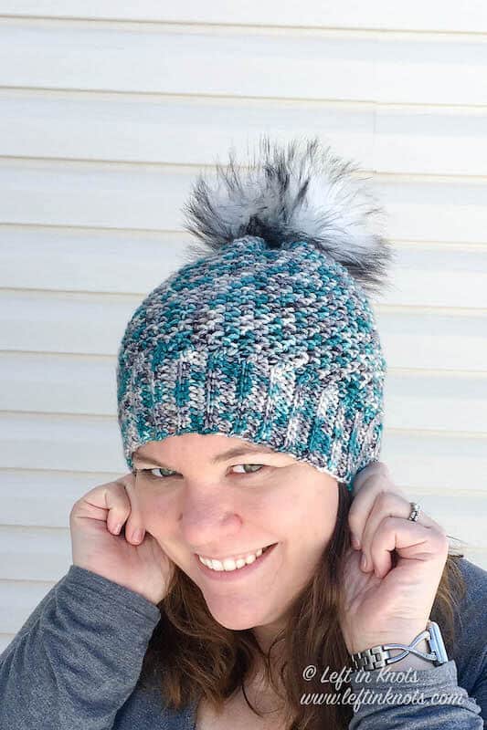 A blue and gray crochet beanie made with hand dyed yarn and a faux fur pom pom