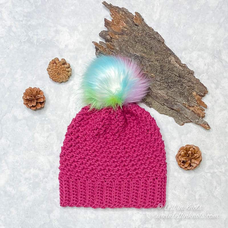 A hot pink crocheted beanie made in a child size with a colorful faux fur pom on top
