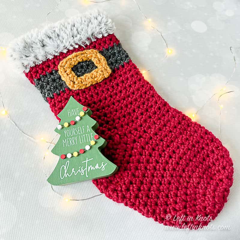A crocheted stocking made with red bulky yarn and a Santa belt. The stocking has a faux fur trim.
