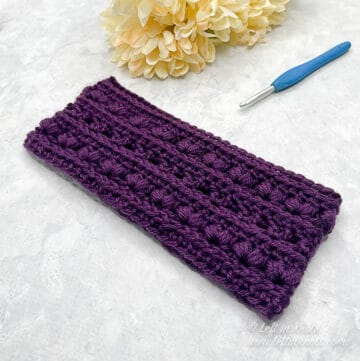 A purple crocheted ear warmer displayed laying next to a crochet hook and ivory colored flowers.
