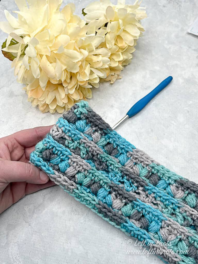 A blue and gray multi colored crochet ear warmer being held and displayed next to ivory flowers and a crochet hook.