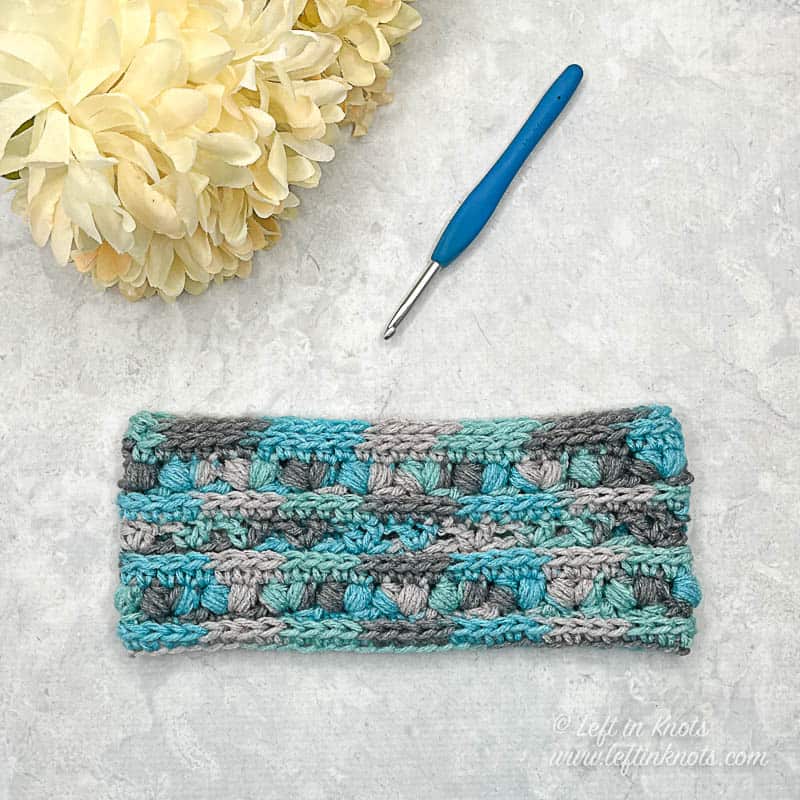 A blue and gray multi colored crochet ear warmer laying flat at displayed next to ivory flowers and a crochet hook.