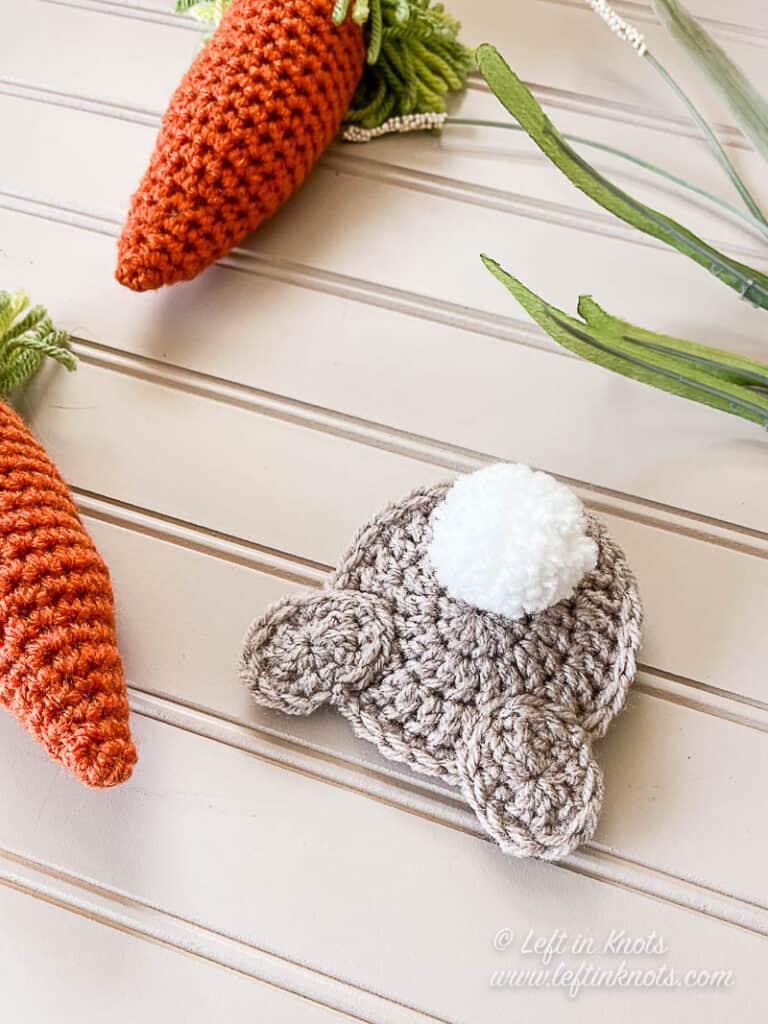 A flat crochet bunny butt applique with a white pom pom tail and hind feet. Displayed next to orange crochet carrots and green grass on a tan background