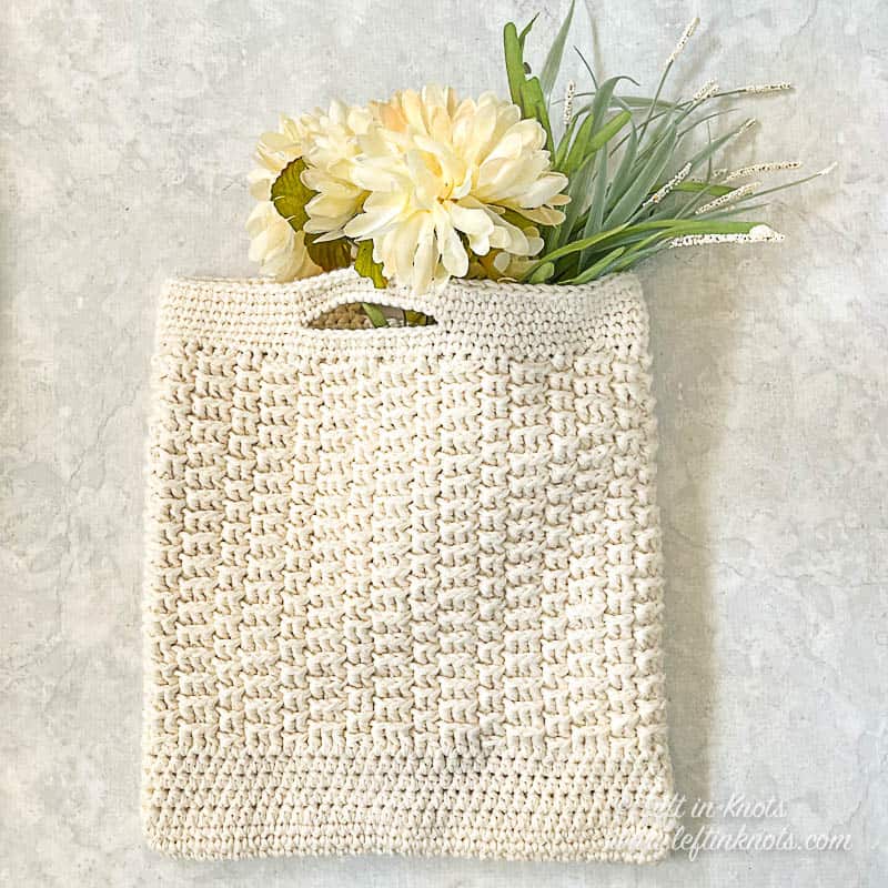A cream colored crochet market bag laying flat. The bag is filled with white flowers and green leaves.