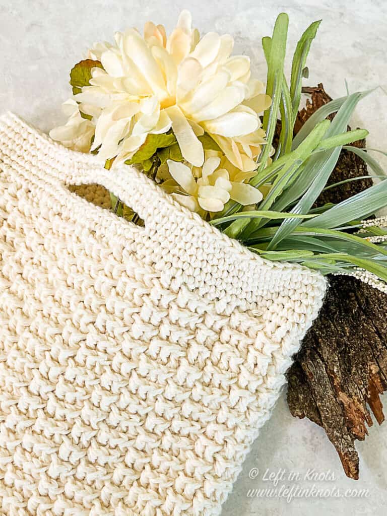 A cream colored crochet market bag laying flat on a piece of tree bark. The bag is filled with white flowers and green leaves.