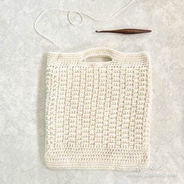 A cream colored crochet market bag laying flat. The bag is displayed next to yarn and a wooden crochet hook.